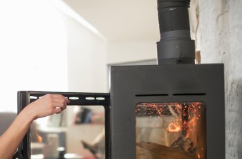 A hand opening the glass door of a wood stove, with glowing embers visible inside, and a vent protruding from the top.