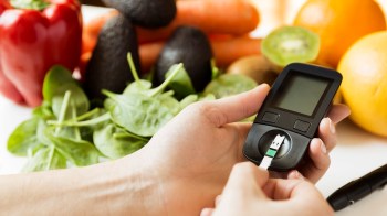 A person using a blood glucose meter with fresh fruits and vegetables in the background.