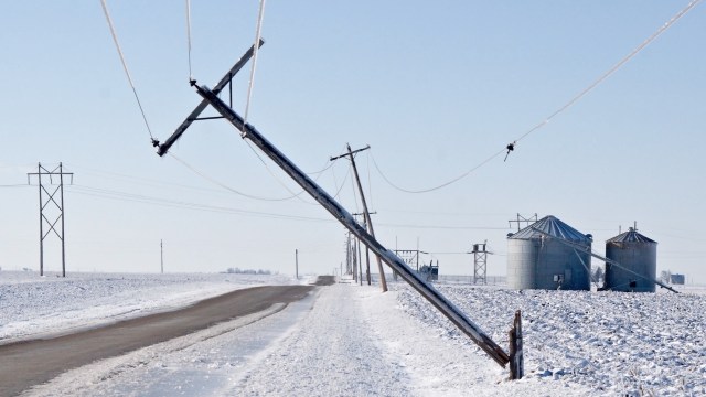 A leaning power pole covered in ice alongside a snowy road and grain silos under a clear sky.
