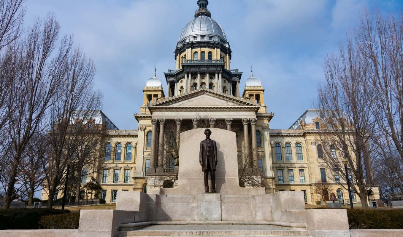 A statue of Abraham Lincoln stands in front of the Illinois Capitol building with a prominent dome under a cloudy sky.