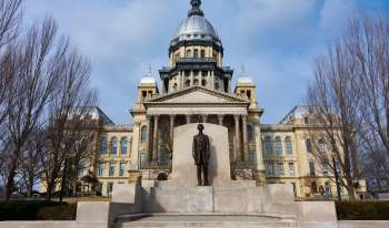 A statue of Abraham Lincoln stands in front of the Illinois Capitol building with a prominent dome under a cloudy sky.
