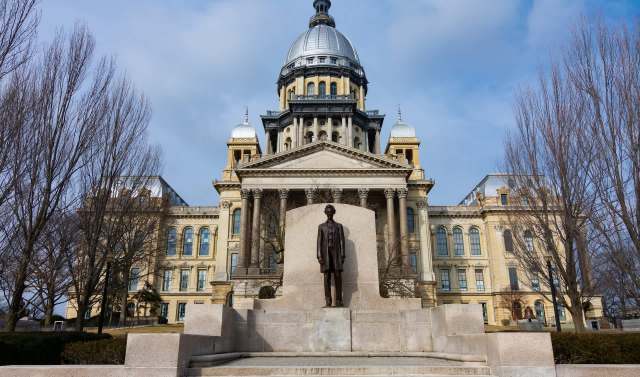 A statue of Abraham Lincoln stands in front of the Illinois Capitol building with a prominent dome under a cloudy sky.