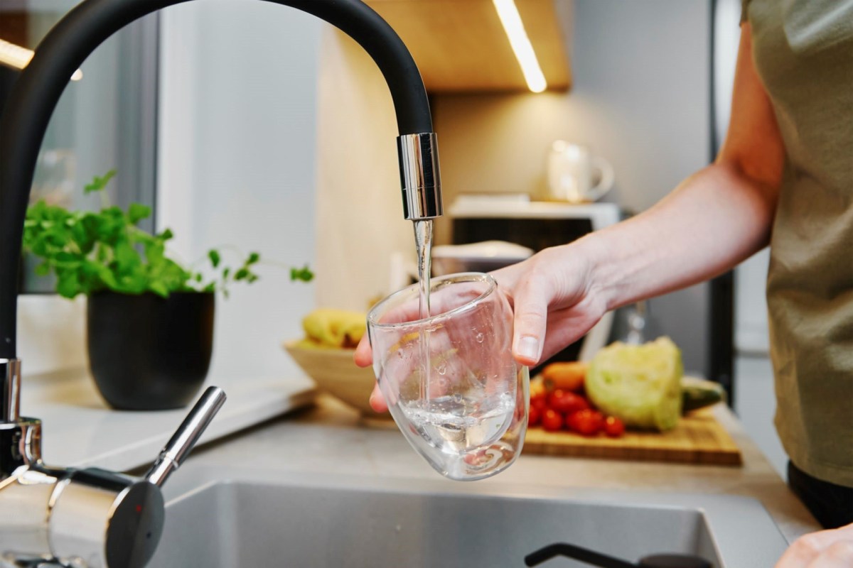 A person fills a clear glass with water from a modern kitchen faucet.