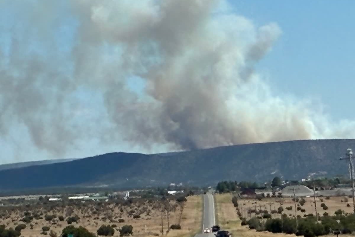 Smoke billows from a hillside as a road leads toward the scene under a clear blue sky.