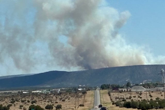 Smoke billows from a hillside as a road leads toward the scene under a clear blue sky.