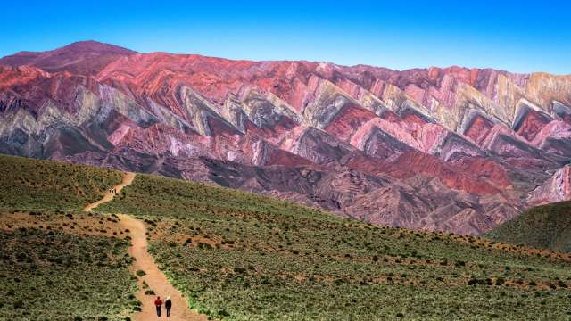 A winding dirt path leads towards colorful, zigzagged mountain formations under a clear blue sky.