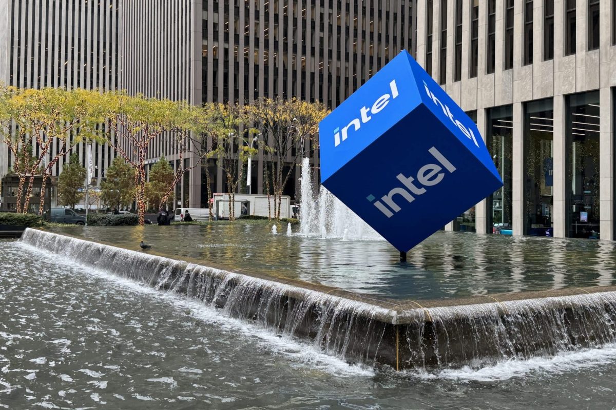 A large blue cube with the Intel logo sits in a water feature surrounded by buildings and trees.