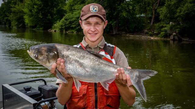 A young man in a life jacket holds a large fish while standing on a boat near a riverbank surrounded by trees.