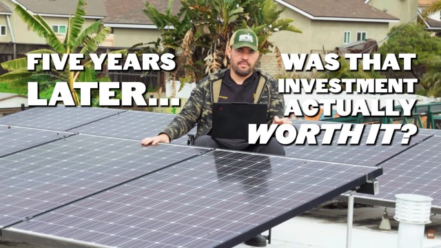 Man sits next to his solar panels with one hand holding a laptop.