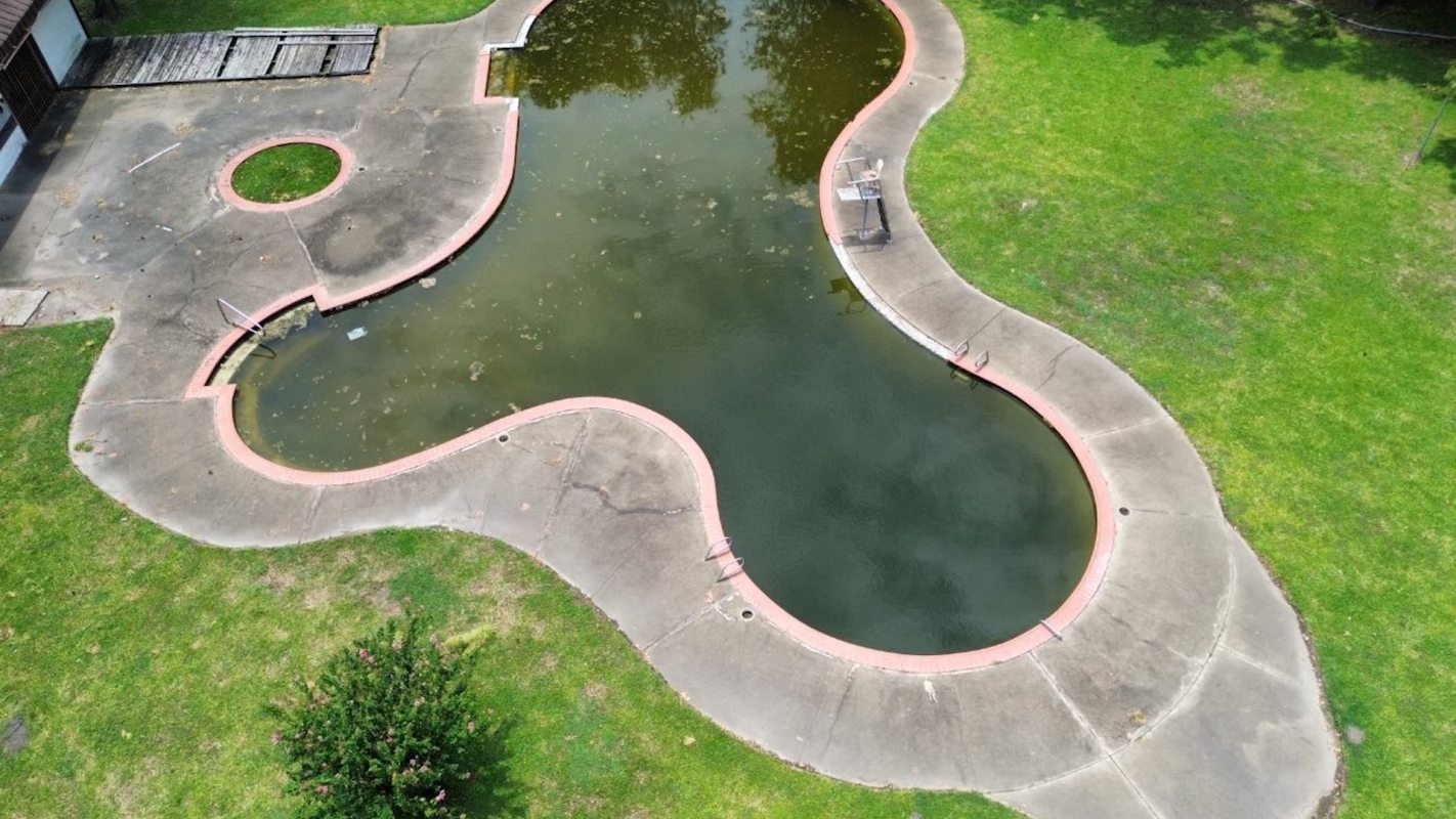 An aerial view of a partially filled, murky swimming pool with a grassy surrounding area.