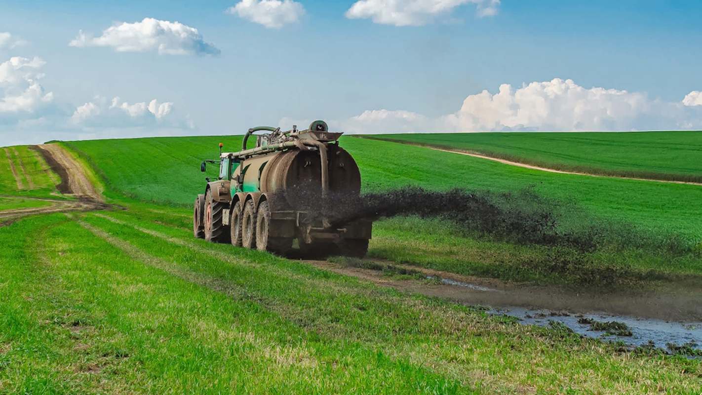 A tractor sprays liquid manure onto a vibrant green field under a blue sky with fluffy clouds.