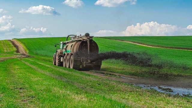 A tractor sprays liquid manure onto a vibrant green field under a blue sky with fluffy clouds.
