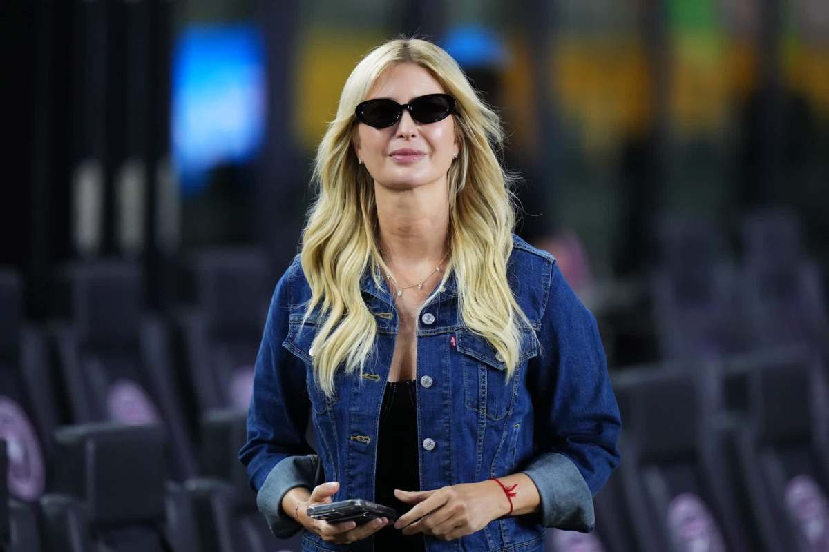 Ivanka Trump wearing sunglasses and a denim jacket, standing in a venue and holding a smartphone.