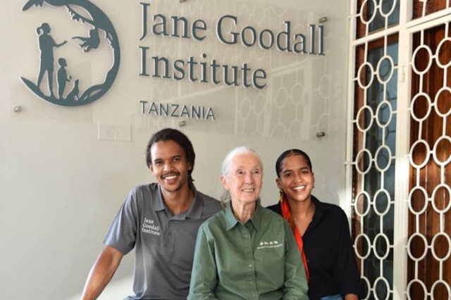 Three individuals pose together in front of the Jane Goodall Institute sign in Tanzania.