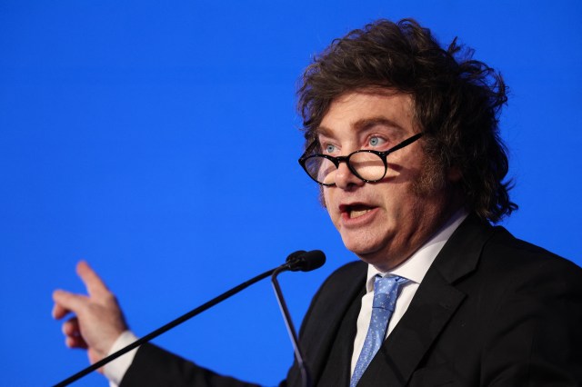 Javier Milei gestures while speaking at a podium against a blue background.