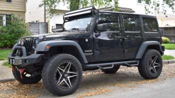 A black Jeep Wrangler with off-road tires and a roof-mounted light bar parked on a suburban street.
