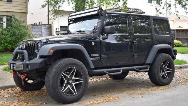 A black Jeep Wrangler with off-road tires and a roof-mounted light bar parked on a suburban street.