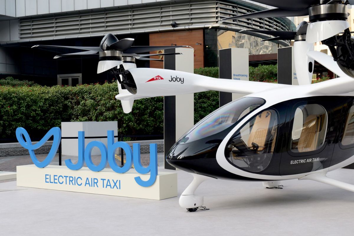 A black and white electric air taxi with a sign displaying "Joby Electric Air Taxi" in a landscaped area.