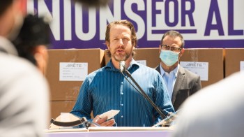 Joe Sanberg speaks at a podium with boxes labeled "ARETE" in the background, while another man listens.