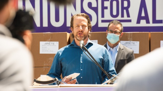 Joe Sanberg speaks at a podium with boxes labeled "ARETE" in the background, while another man listens.
