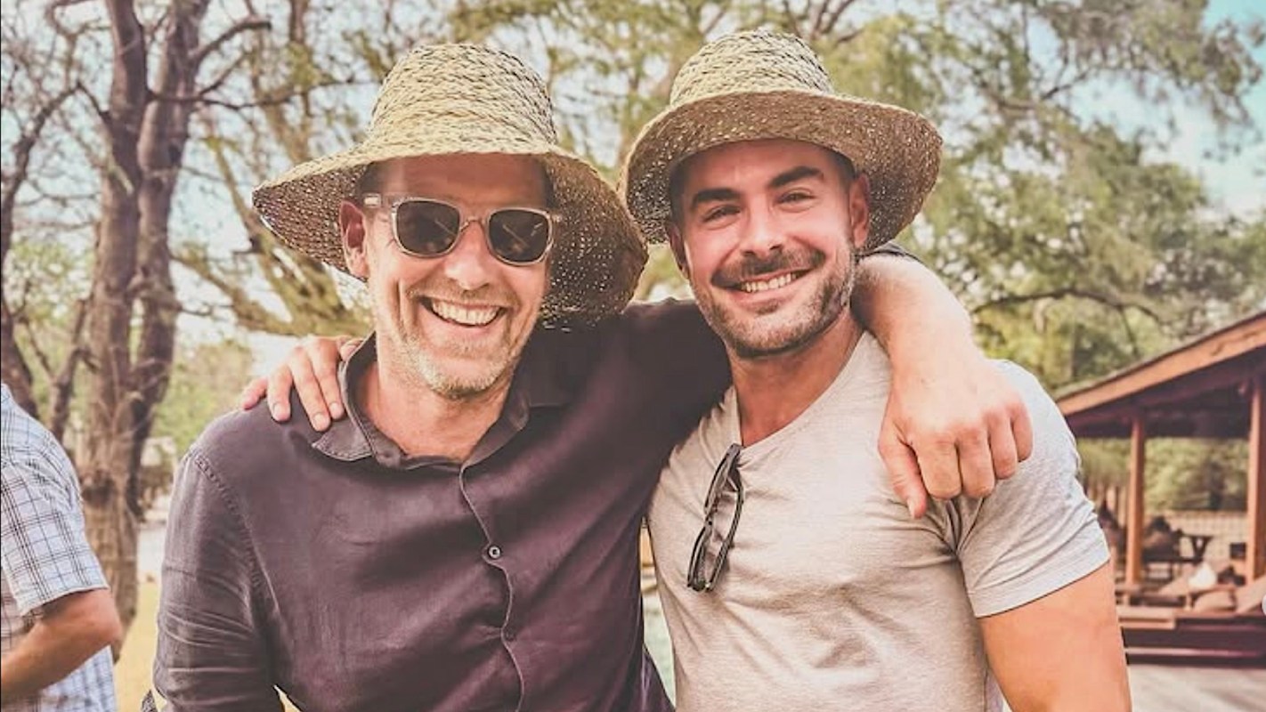 Joost Bakker and Zac Efron smile and pose together outdoors, embracing under sunlight with trees in the background.