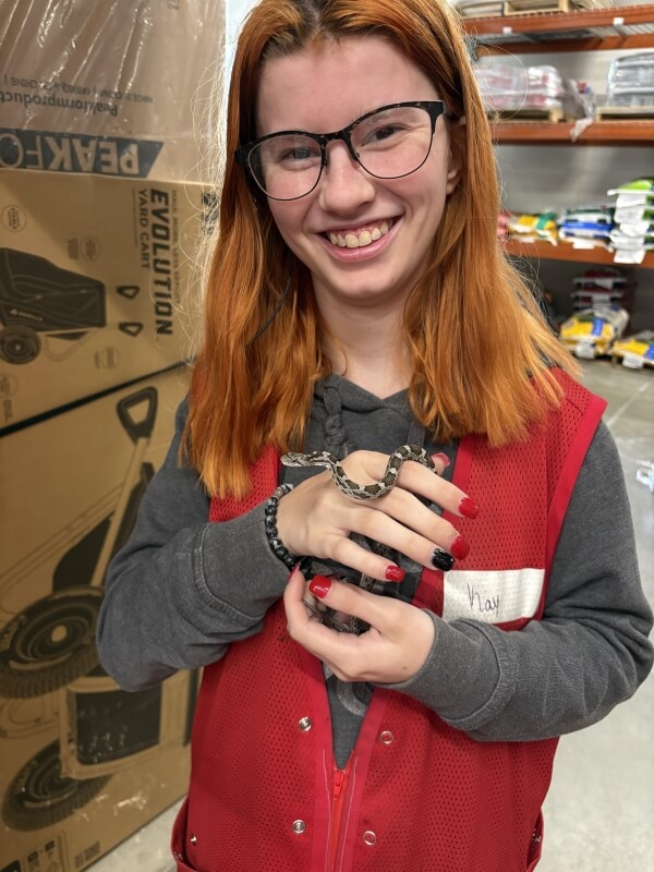 A person with red and black nails holds a small snake, wearing a gray sweatshirt and a red vest with a name tag.