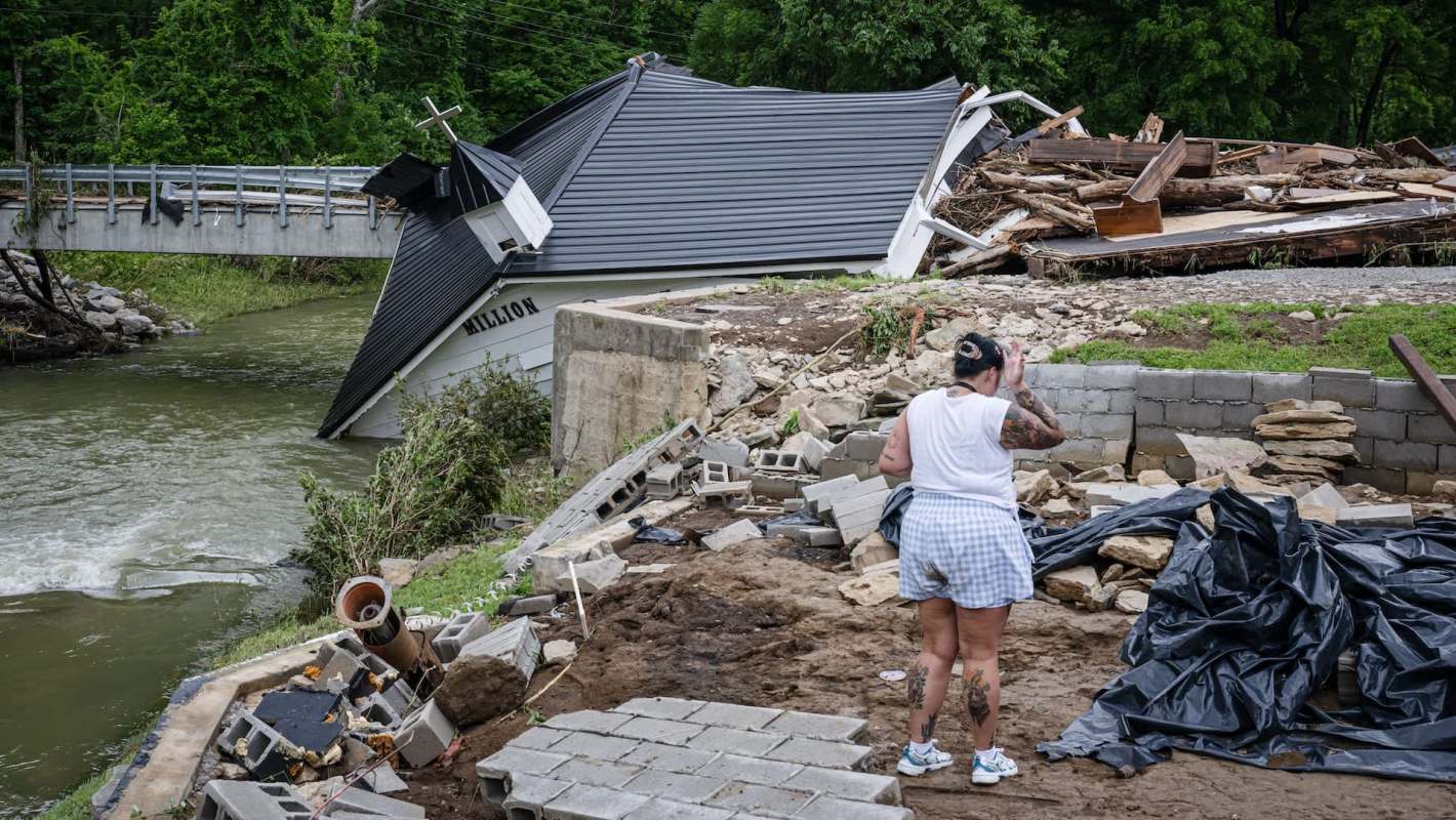 A woman stands amid debris near a flooded building partially collapsed by a riverbank.