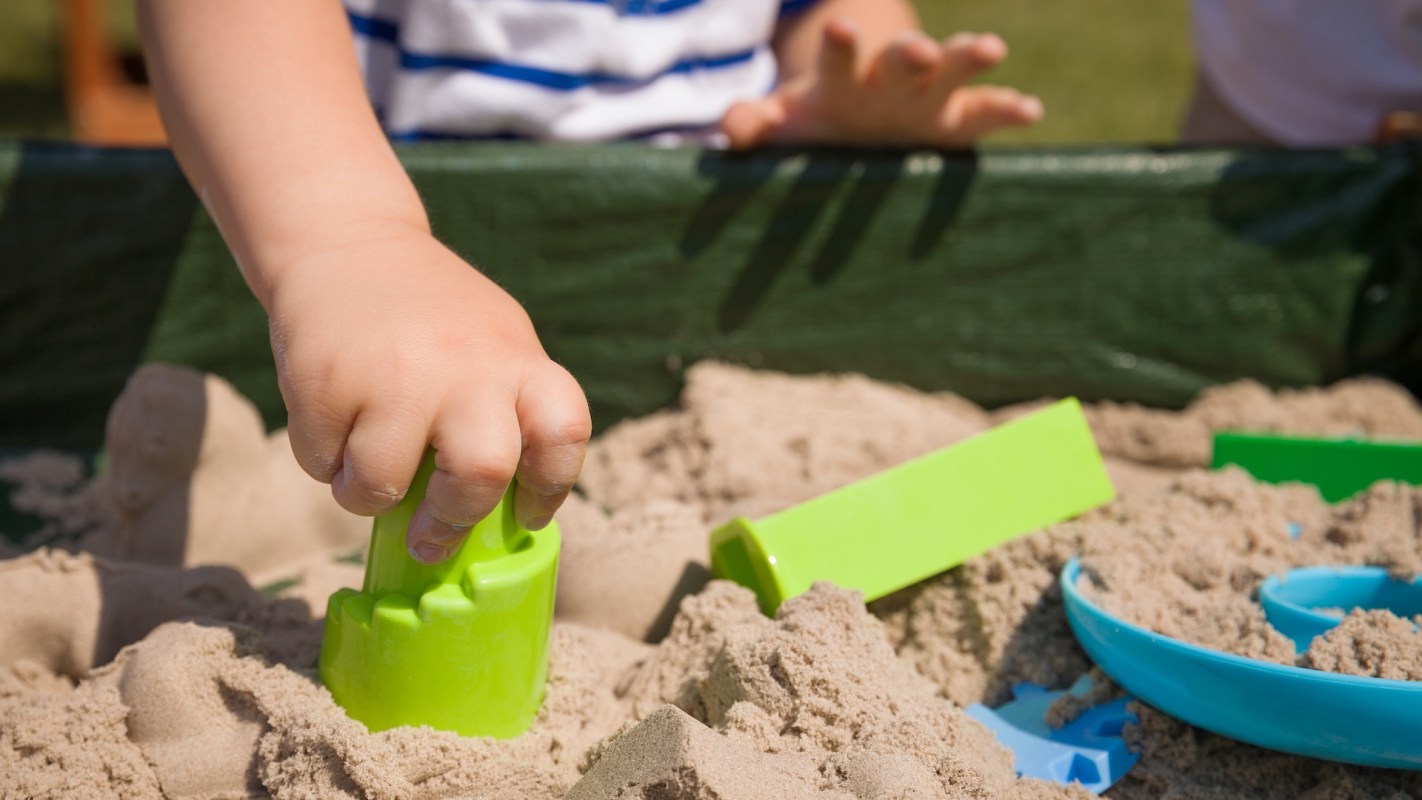A child's hand grips a green sand mold while playing in a sandbox surrounded by additional colorful toys.