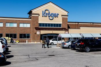 A Kroger grocery store front with shoppers, parked cars, and stacked bags outside on a sunny day.