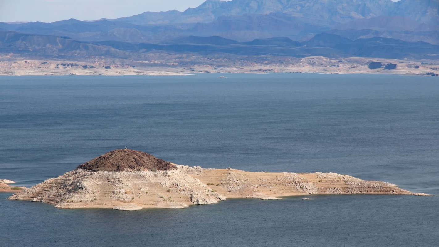 A small island with rocky terrain and sparse vegetation surrounded by calm blue water and distant mountain ranges.