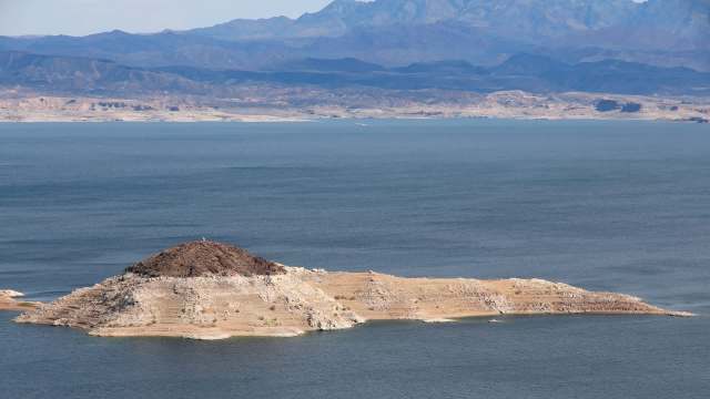 A small island with rocky terrain and sparse vegetation surrounded by calm blue water and distant mountain ranges.