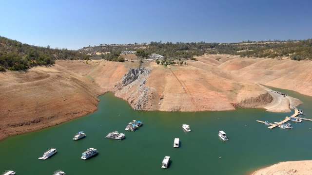 Aerial view of a dry landscape with boats docked in water surrounded by red hills under a clear blue sky.