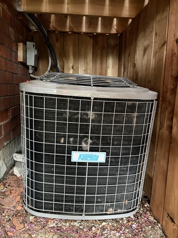 An outdoor air conditioning unit placed against a wooden wall and adjacent to a brick structure.