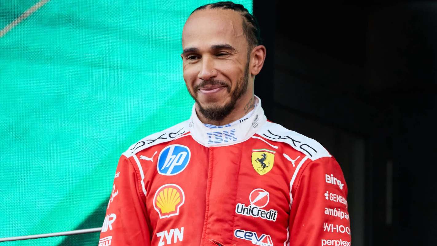 Lewis Hamilton stands in front of a green backdrop wearing a red racing suit, showcasing sponsor logos.
