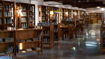 A spacious library with wooden bookshelves, tables, and chairs, softly lit for a cozy reading atmosphere.