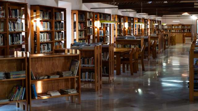 A spacious library with wooden bookshelves, tables, and chairs, softly lit for a cozy reading atmosphere.