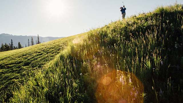 A golfer swings a club on a grassy hillside under a bright sun, with mountains in the background.