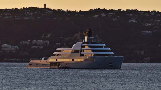 A large luxury yacht anchored in calm waters with hills and buildings in the background during sunset.