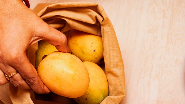 A hand reaches into a cloth bag, grasping a ripe mango among several others.