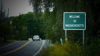 A green welcome sign indicating entry into Massachusetts along a winding road surrounded by trees.