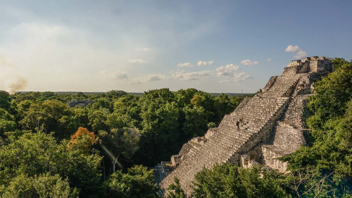 A person climbs the steps of a large ancient pyramid surrounded by lush green trees under a clear sky.