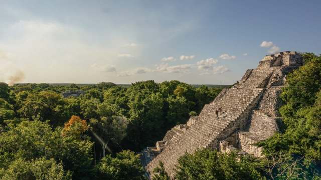 A person climbs the steps of a large ancient pyramid surrounded by lush green trees under a clear sky.