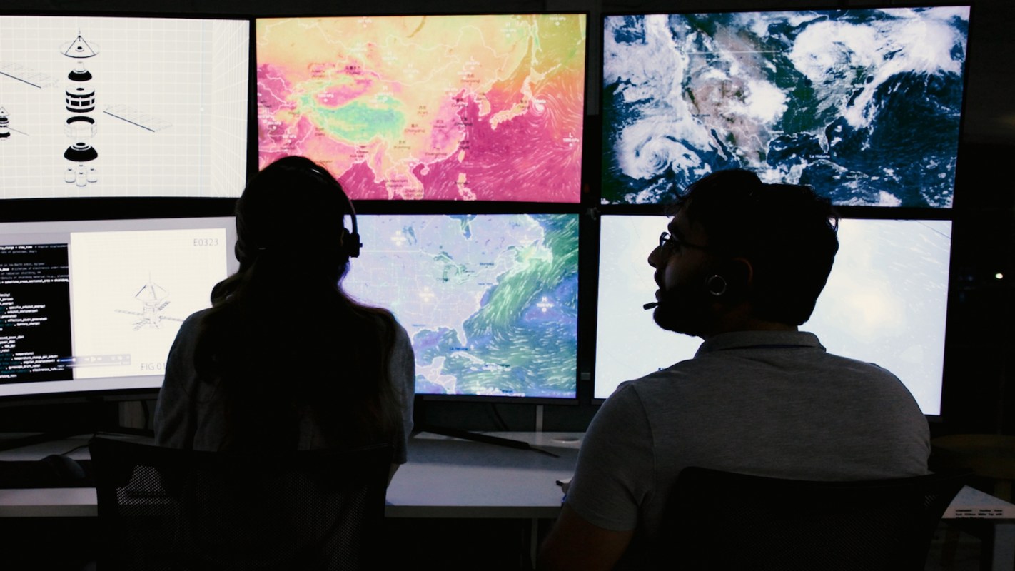 Two individuals with headsets observe screens satellite images.