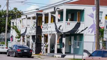 A colorful two-story building with abstract murals, balconies, and parked cars on a sunny street.