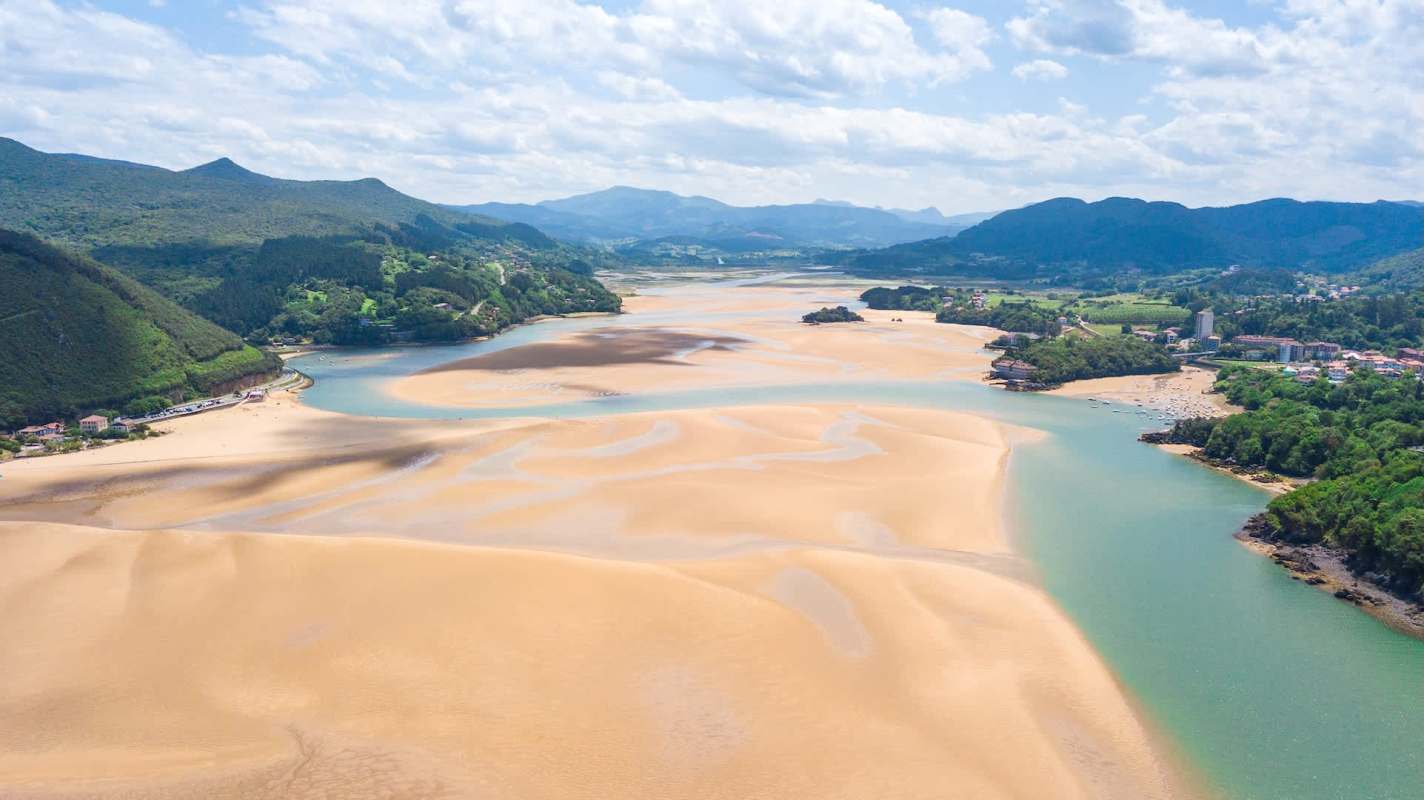 Aerial view of a river winding through sandy banks under a partly cloudy sky, surrounded by lush green hills.
