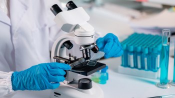 A researcher in blue gloves places a slide under a microscope in a lab filled with test tubes and glassware.