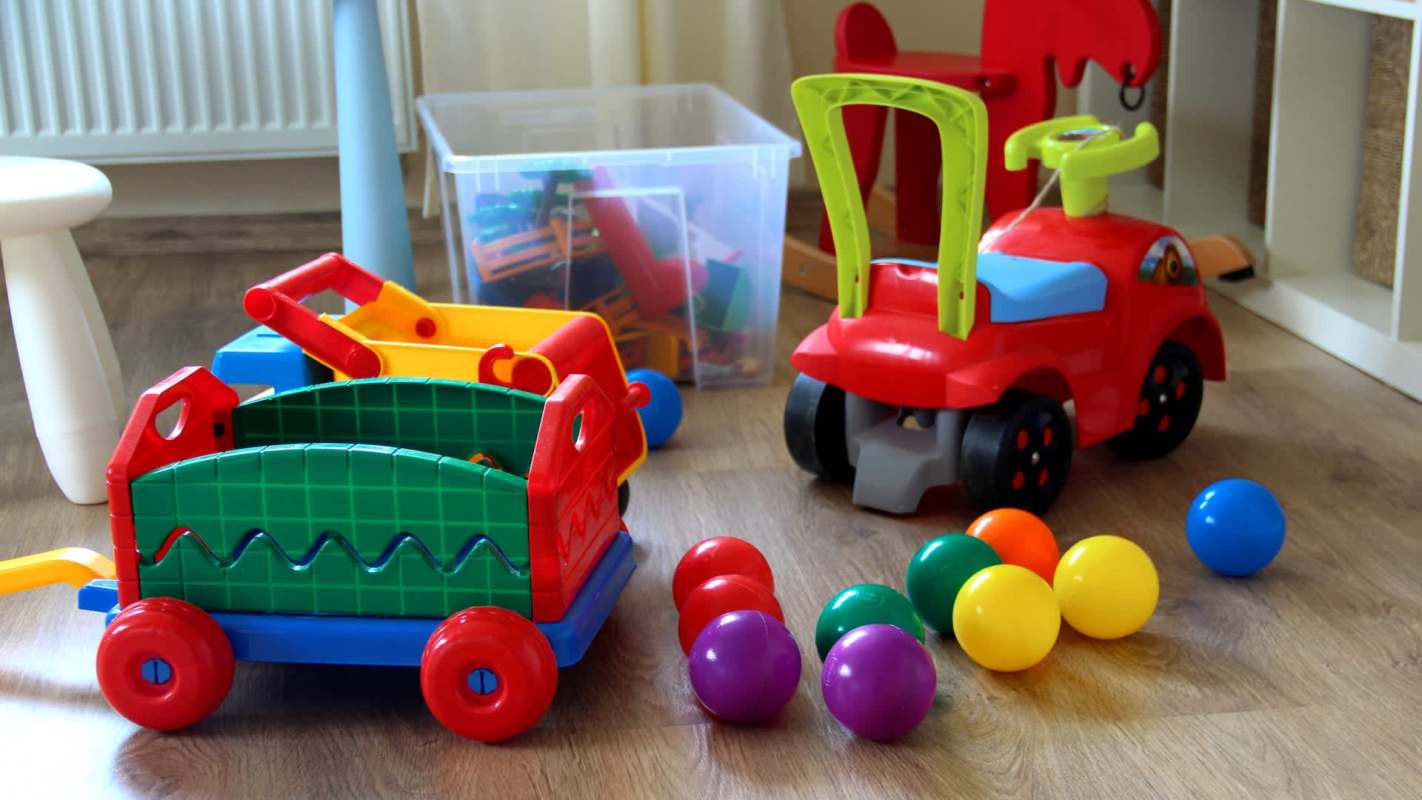 A colorful array of plastic toy vehicles, carts, and balls scattered across a wooden floor.