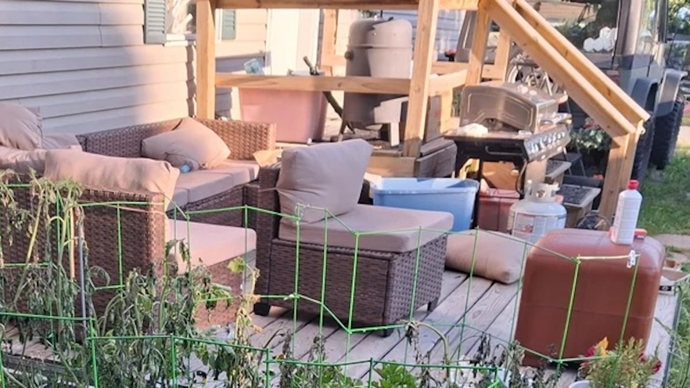 A cluttered outdoor patio with wicker furniture, a grill, and pots among wilting plants.