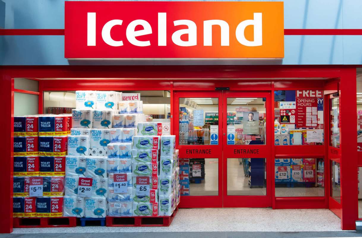An Iceland store entrance featuring stacks of toilet paper and kitchen rolls outside.