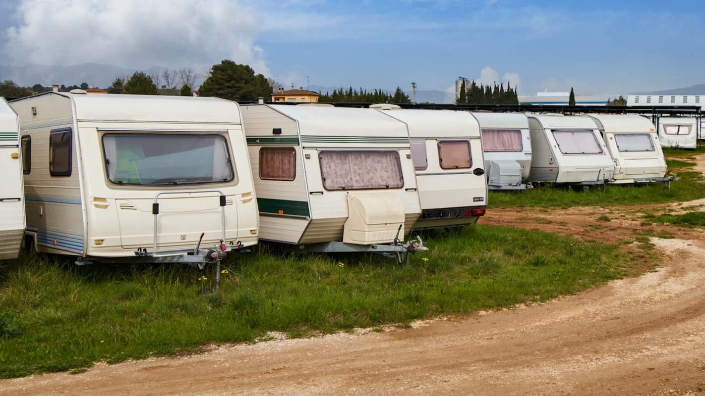 A row of parked caravans on a grassy area beside a dirt path under a partly cloudy sky.